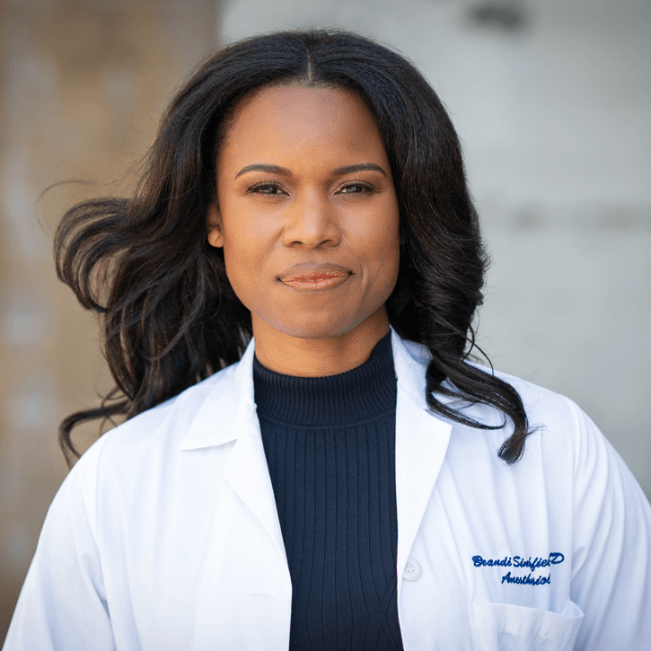 An image a Dr. Brandi, an adult Black woman with long dark hair, smirking into the camera. She is wearing a white lab coat.