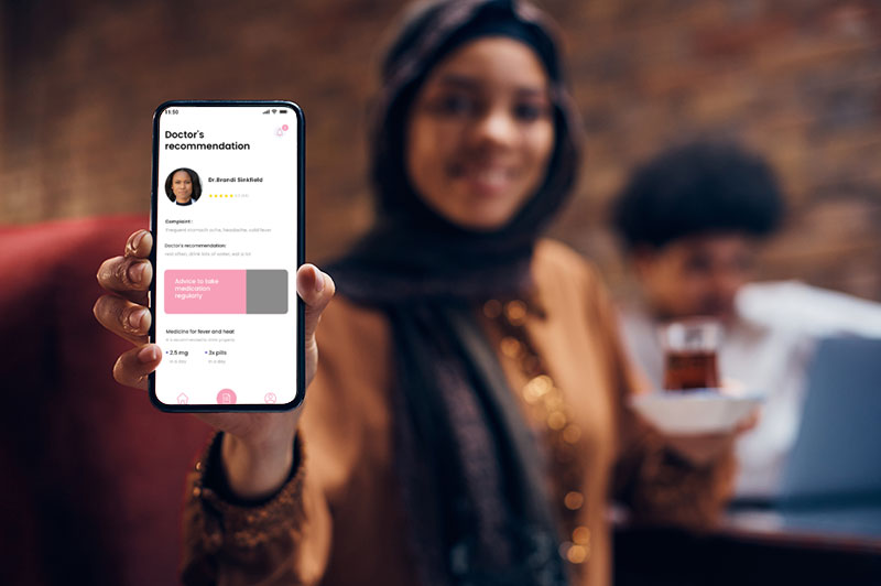 A woman wearing a headscarf is holding a cell phone with the screen facing outward. On the screen is a small image of a health app.